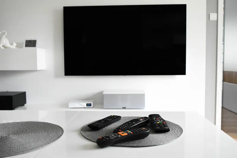 Televisão desligada na parede de uma sala branca, com controles remotos sobre a mesa