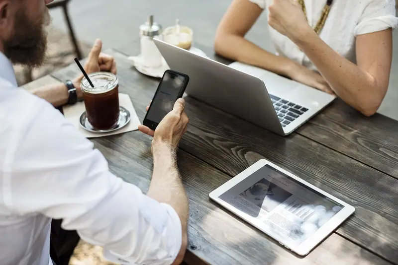 Mãos de pessoas numa mesa de madeira com notebook, tablet, celular e um copo de café
