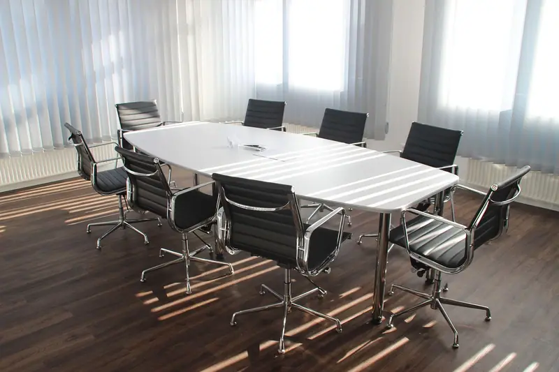 Sala de reunião vazia com mesa branca, cadeiras pretas e luz do sol entrando pelas cortinas