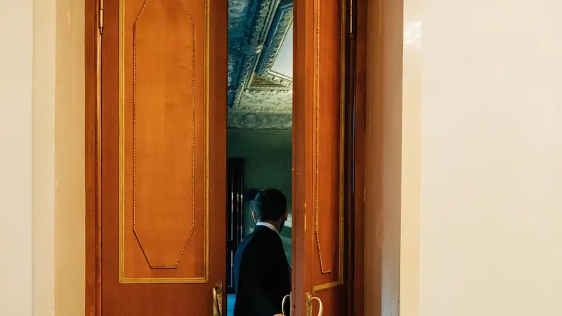 Pessoa de costas abrindo uma porta alta de madeira envernizada, com outra sala ao fundo