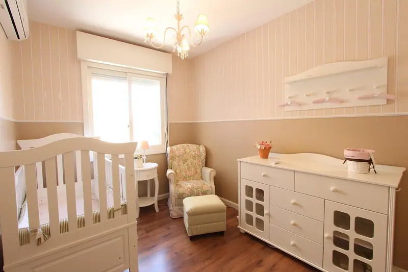Quarto de bebê claro com berço, poltrona, cômoda, janela e aparelho de ar-condicionado