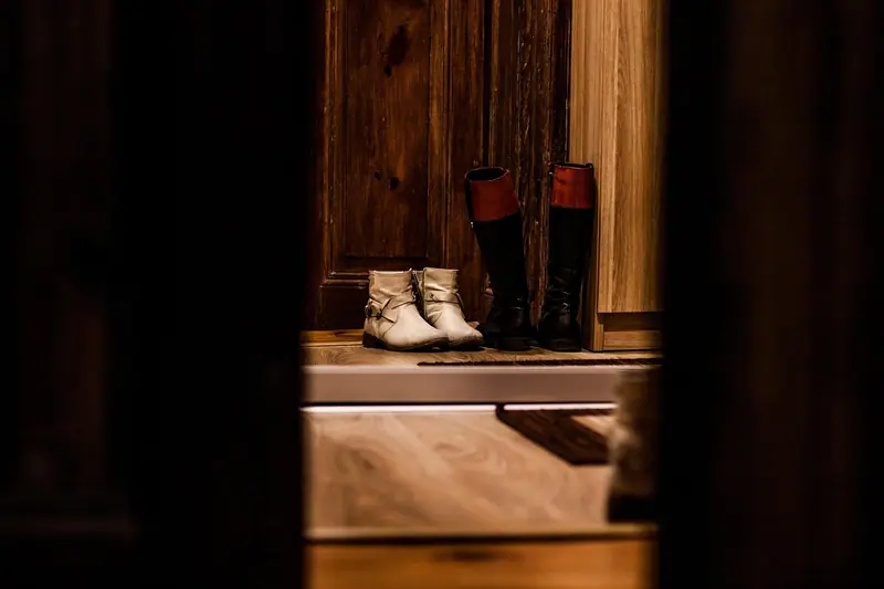 Botas e sapatos no chão junto a uma porta de madeira, vistos pela fresta de outra porta