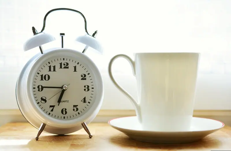 Relógio despertador branco ao lado de uma xícara de café sobre a mesa