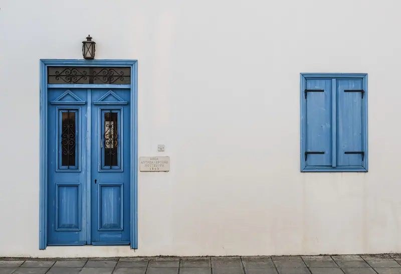 Porta dupla azul com vidros e bandeira no alto, ao lado de uma janela azul numa fachada branca
