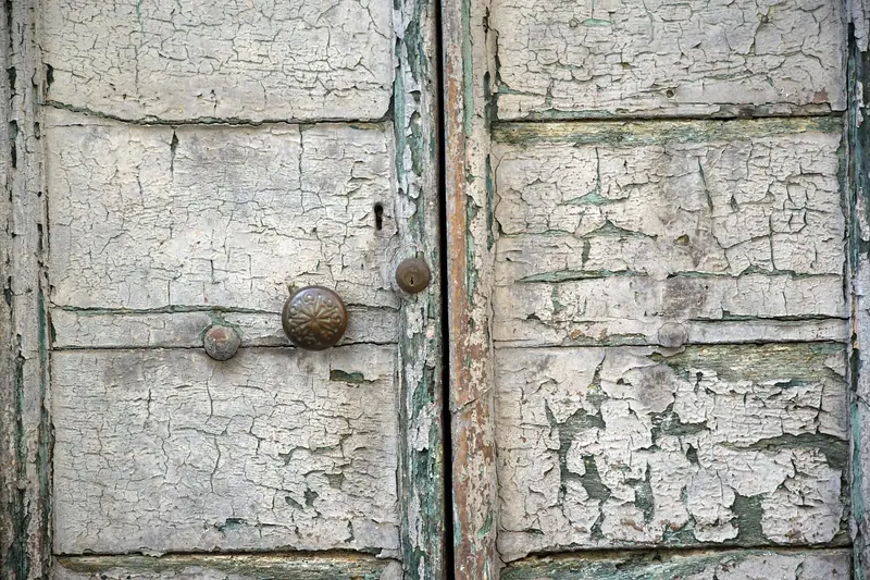 Porta dupla de madeira com a pintura branca rachada e descascando, e maçanetas redondas de metal
