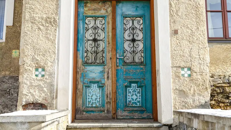 Porta dupla de madeira azul com grades decoradas nos vidros, numa fachada clara com degrau de pedra