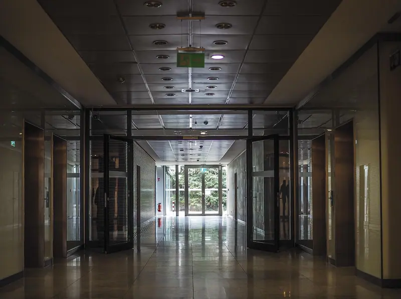 Corredor de prédio comercial com duas portas de vidro de abrir, de moldura escura, abertas para os lados