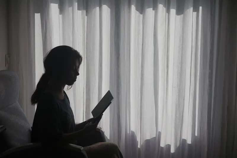 Silhueta de uma mulher lendo um livro ao lado de uma janela com cortina clara