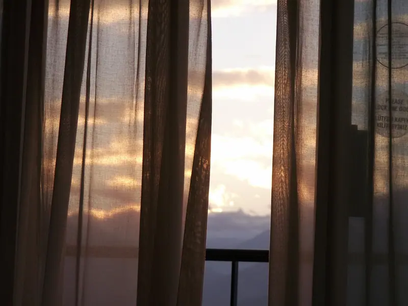 Cortinas finas diante de uma janela de vidro, com a luz do amanhecer e o guarda-corpo da varanda
