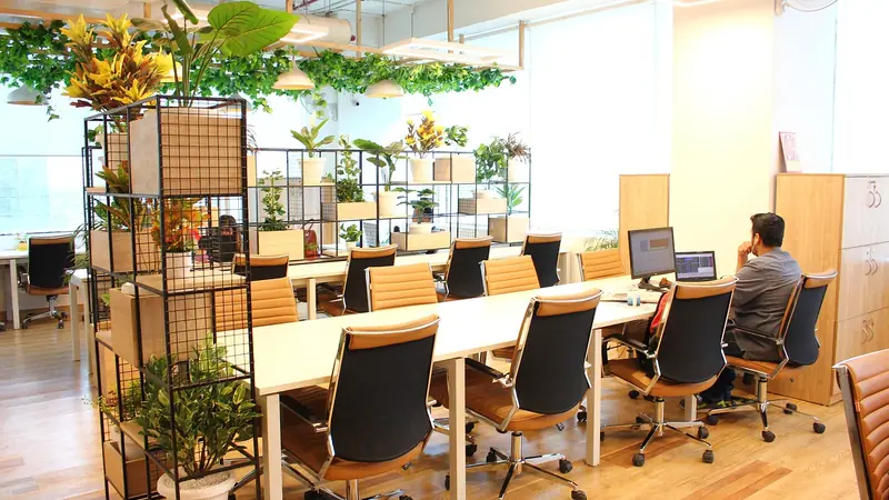 Espaço de coworking com mesas compartilhadas, cadeiras de couro, estantes com plantas e uma pessoa trabalhando ao fundo