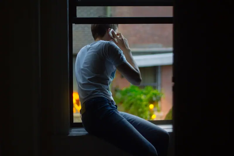 Homem de costas falando ao celular, sentado no parapeito de uma janela, num ambiente escuro