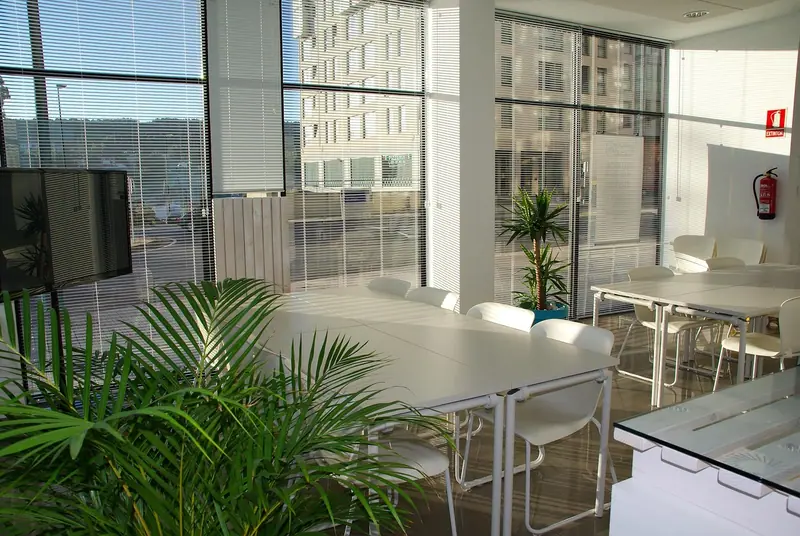 Sala de trabalho com mesas brancas, persianas e janelas de vidro do piso ao teto voltadas para a rua