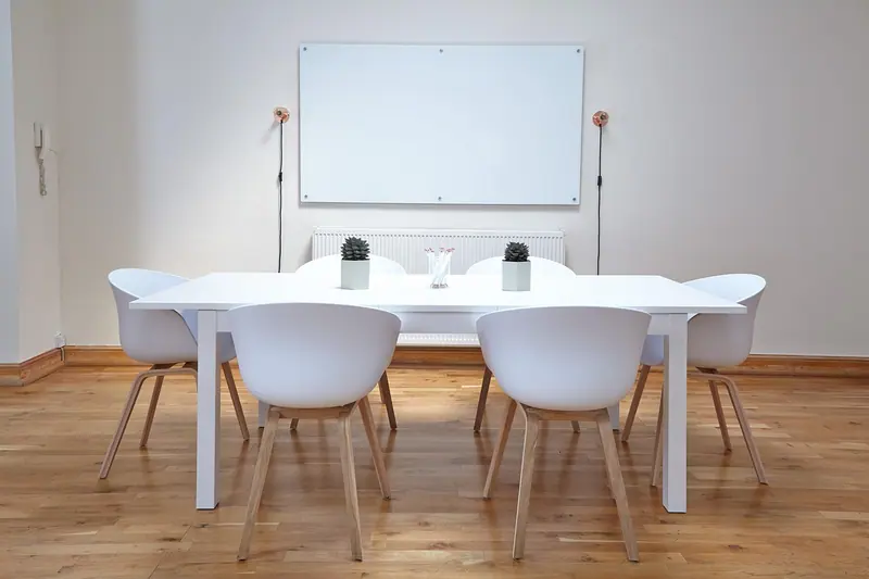 Sala de reunião pequena com mesa branca, cadeiras brancas, quadro branco na parede e piso de madeira