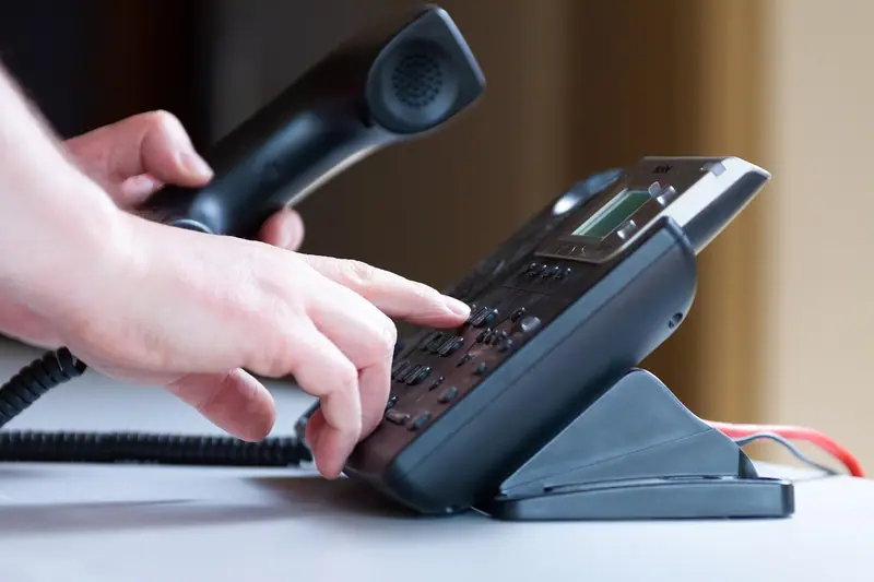 Mão segurando o fone de um telefone de mesa enquanto a outra digita no teclado