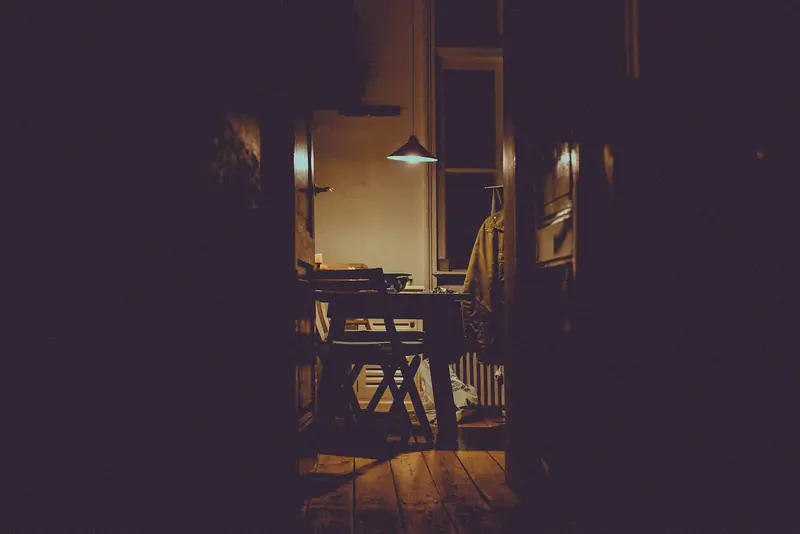 Interior de uma casa à noite, com uma luminária acesa sobre a mesa e o resto na penumbra