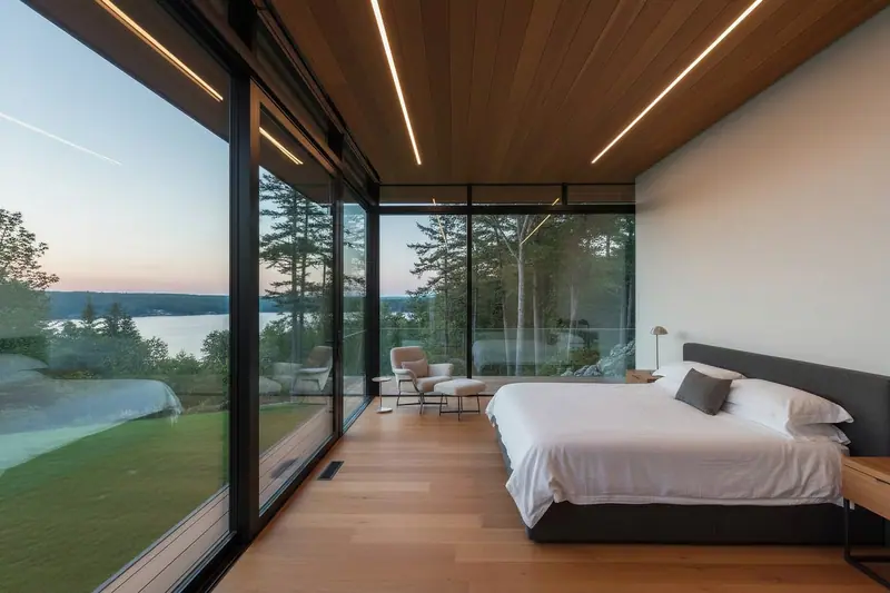 Quarto com cama de casal e uma parede inteira de vidro com vista para árvores e um lago