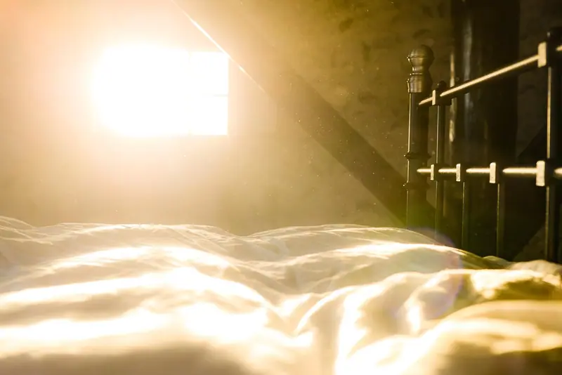 Cama com lençóis claros iluminada pela luz do sol que entra por uma janela