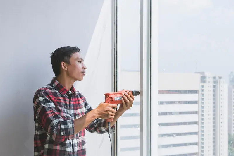 Homem segurando uma furadeira ao lado de uma janela grande, com prédios ao fundo