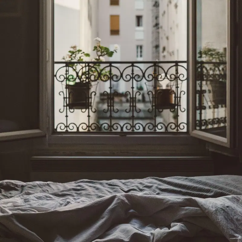 Cama desarrumada diante de uma janela de abrir com as folhas abertas e um guarda-corpo de ferro