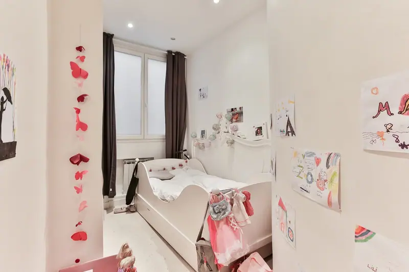 Quarto infantil claro, com cama branca, desenhos na parede e janela ao fundo