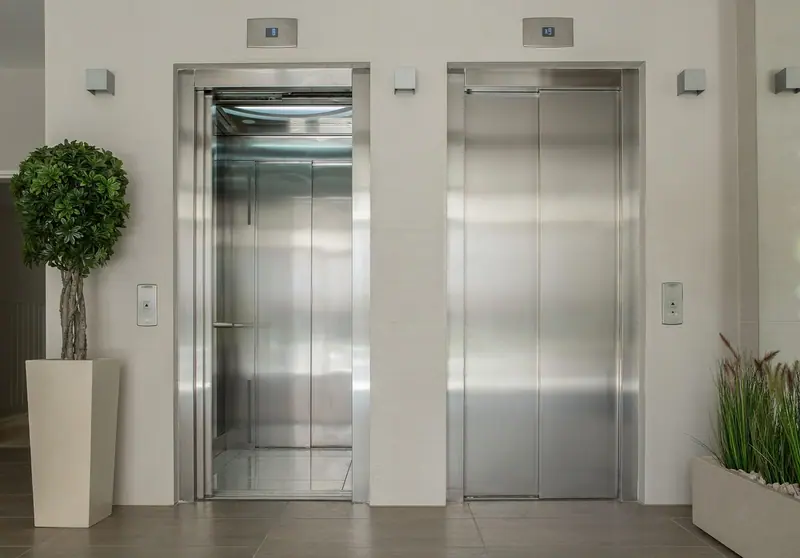 Hall de prédio com duas portas de elevador de inox, vasos de plantas e piso claro
