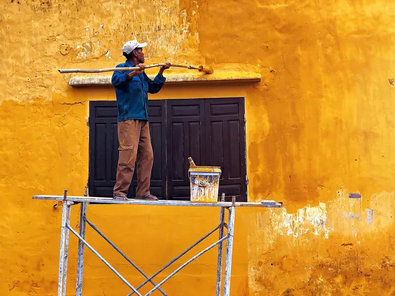Pintor de costas sobre um andaime, pintando uma parede amarela acima de uma janela fechada