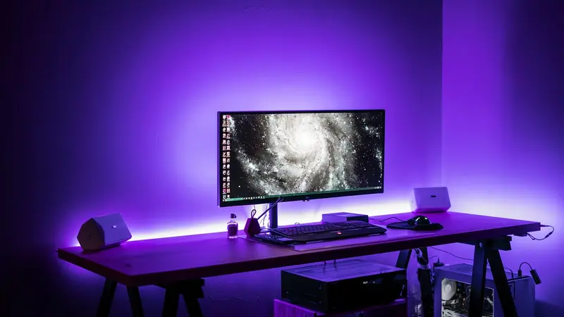 Mesa com monitor e computador iluminada por luz roxa contra a parede