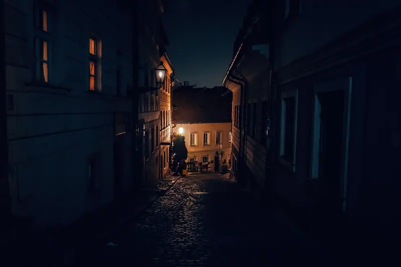 Rua estreita à noite, entre prédios, iluminada por um poste ao fundo