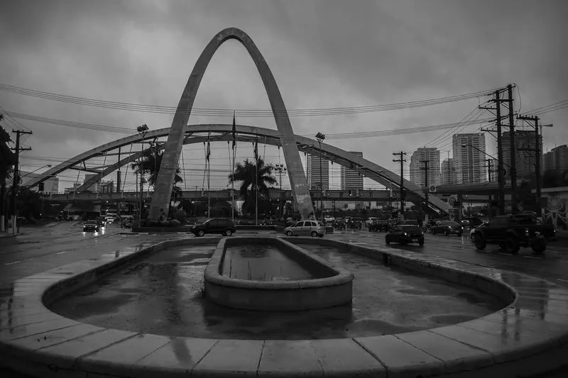 Cruzamento com carros, postes e prédios ao fundo, sob um grande arco de concreto, em preto e branco