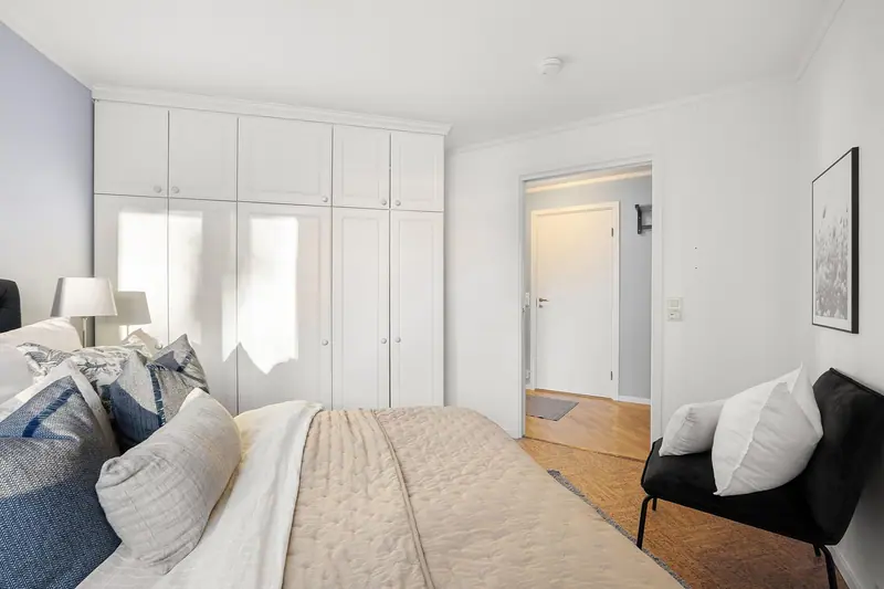 Quarto claro com cama de casal arrumada, almofadas, poltrona preta e armário branco
