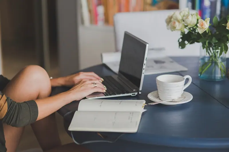 Pessoa trabalhando num notebook numa mesa com xícara de café, caderno e flores