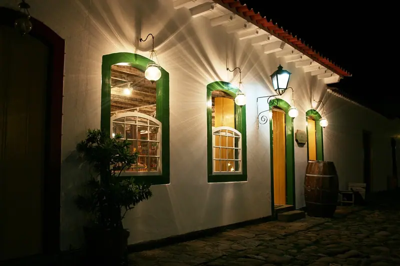 Fachada de casa antiga à noite, com janelas verdes iluminadas por luminárias na rua de pedra