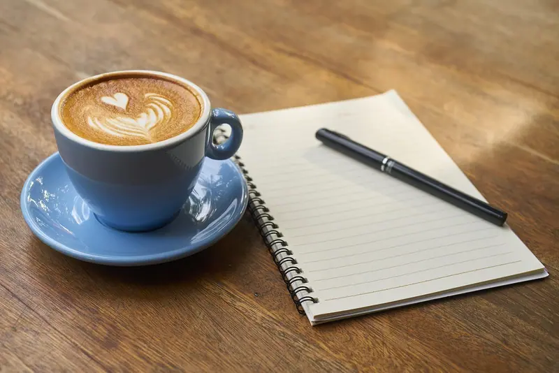 Caderno aberto com uma caneta preta sobre uma mesa de madeira, ao lado de uma xícara de café