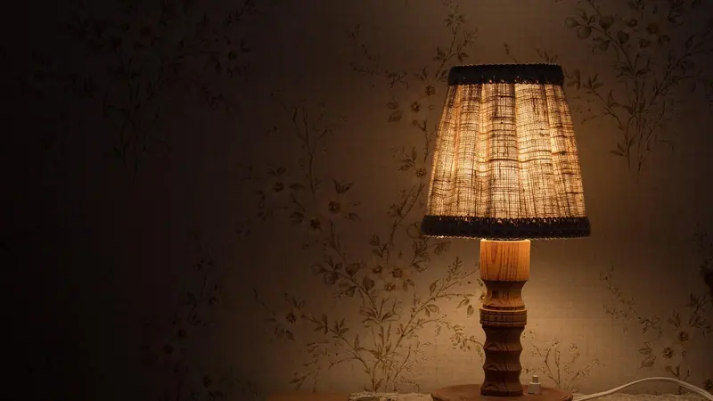Abajur com cúpula de tecido aceso sobre uma mesa de cabeceira, num quarto à meia-luz