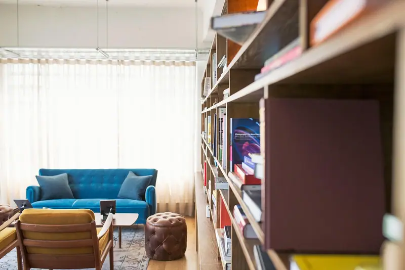 Sala clara com sofá azul, poltronas e uma estante de livros diante de janelas com cortinas brancas