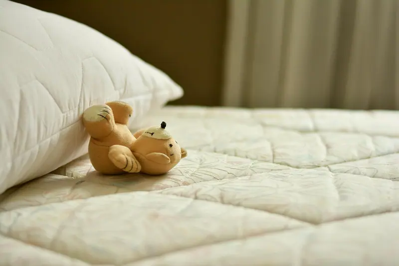 Pequeno urso de brinquedo deitado sobre um colchão claro, ao lado do travesseiro