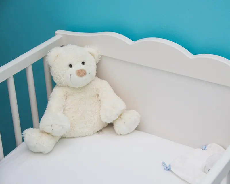 Urso de pelúcia branco sentado dentro de um berço branco, com parede azul ao fundo