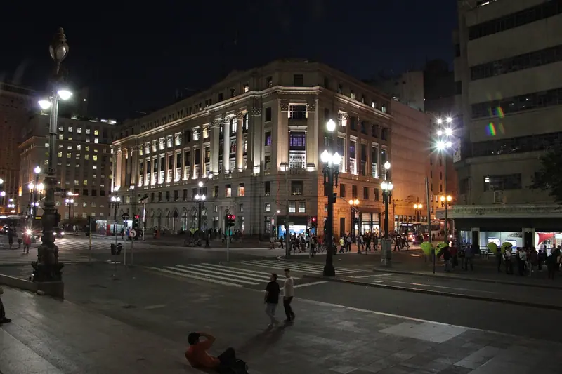 Praça do centro de São Paulo à noite, com prédio histórico iluminado e poucas pessoas ao longe