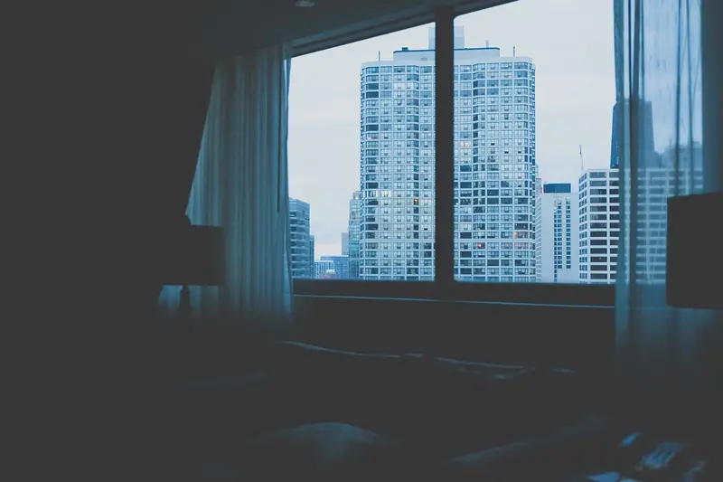 Quarto em penumbra com cortina aberta e janela voltada para prédios altos