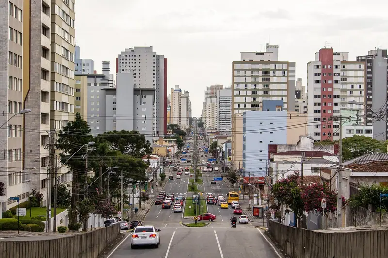 Avenida com carros e ônibus subindo entre prédios residenciais numa cidade brasileira