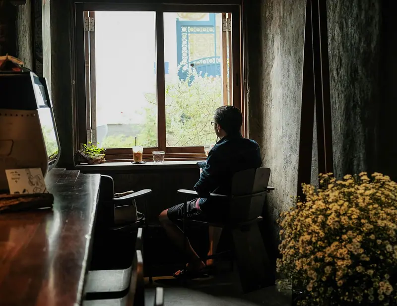 Homem sentado de costas diante de uma janela de madeira, num ambiente com flores amarelas