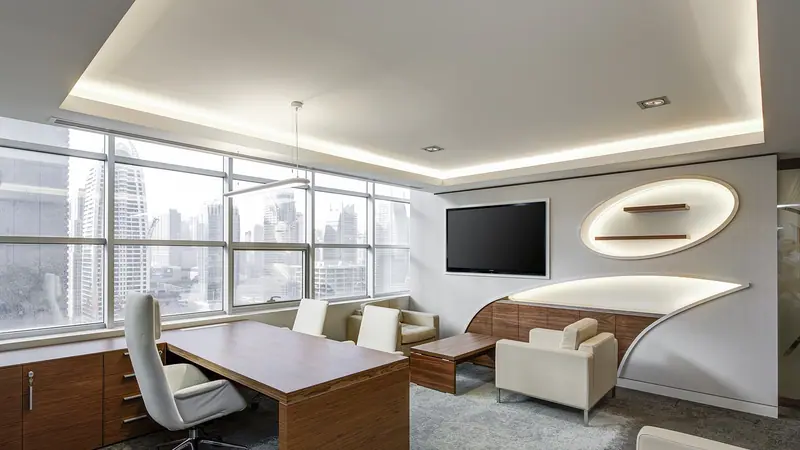 Sala executiva com mesa de madeira, cadeira branca, sofá e janelas amplas com vista para prédios