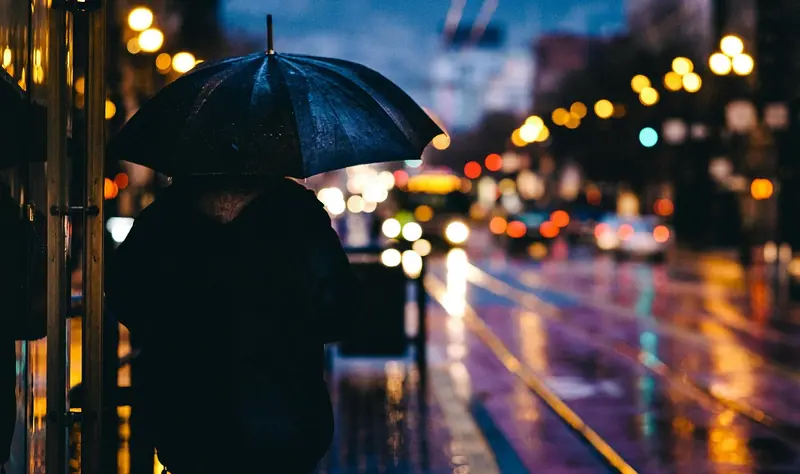 Pessoa de costas segurando um guarda-chuva numa rua molhada, com luzes da cidade desfocadas à noite