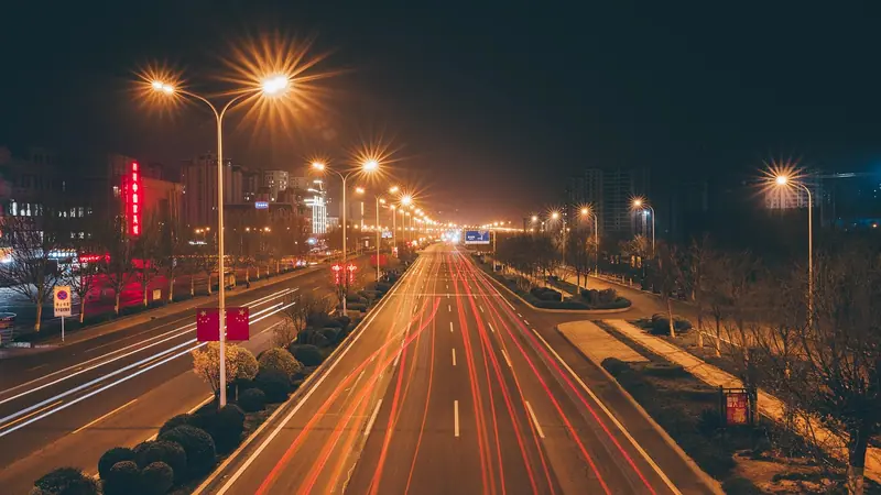 Avenida larga à noite, com postes acesos e rastros de luz dos carros nas duas pistas