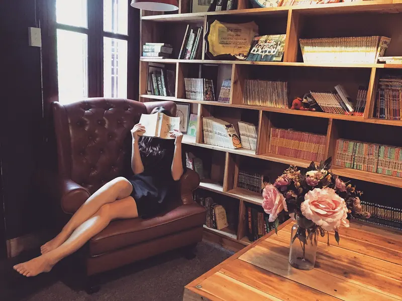 Mulher lendo numa poltrona de couro, com o livro cobrindo o rosto, ao lado de uma estante e de um vaso de flores