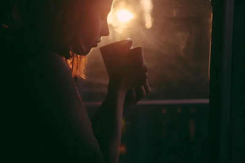 Silhueta de uma mulher tomando algo numa caneca, junto a uma janela com luz quente do fim de tarde