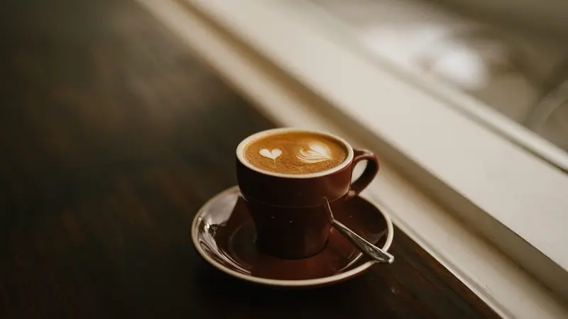 Xícara de café com desenho na espuma sobre o pires, numa mesa de madeira junto a uma janela clara
