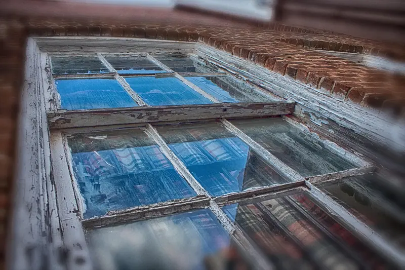 Janela antiga de caixilho branco vista de perto e de lado, com os vidros refletindo o céu