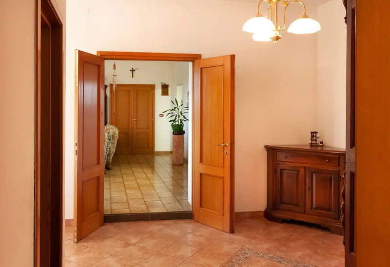 Porta de madeira aberta para um hall de piso cerâmico, com aparador e lustre