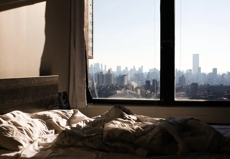 Quarto com cama desarrumada diante de uma janela grande com prédios ao fundo